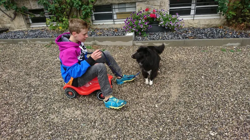 Ein junger Junge sitzt auf einem Scoote mit einem Hund. – Jugend und Hund Lager 2017 in Balmberg