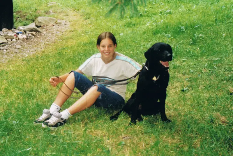 Eine Frau im Gras mit ihrem schwarzen Hund, trägt ein weisses und blaues Kleid und lächelnd. Rasse: Labrador Retriever. – Jugend und Hund Lager 2001 in Eigenthal