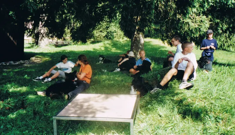 Eine Gruppe von Menschen, die auf einem üppigen grünen Feld sitzen, mit zwei Hunden im Vordergrund. – Jugend und Hund Lager 2001 in Eigenthal
