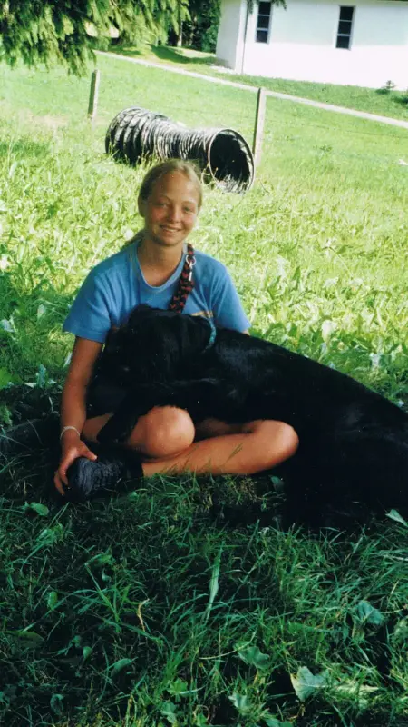 Eine Frau, die mit einem schwarzen Hund im Gras sitzt, ein blaues T-Shirt trägt und lächelt. Rasse: Riesenschnauzer. – Jugend und Hund Lager 2001 in Eigenthal