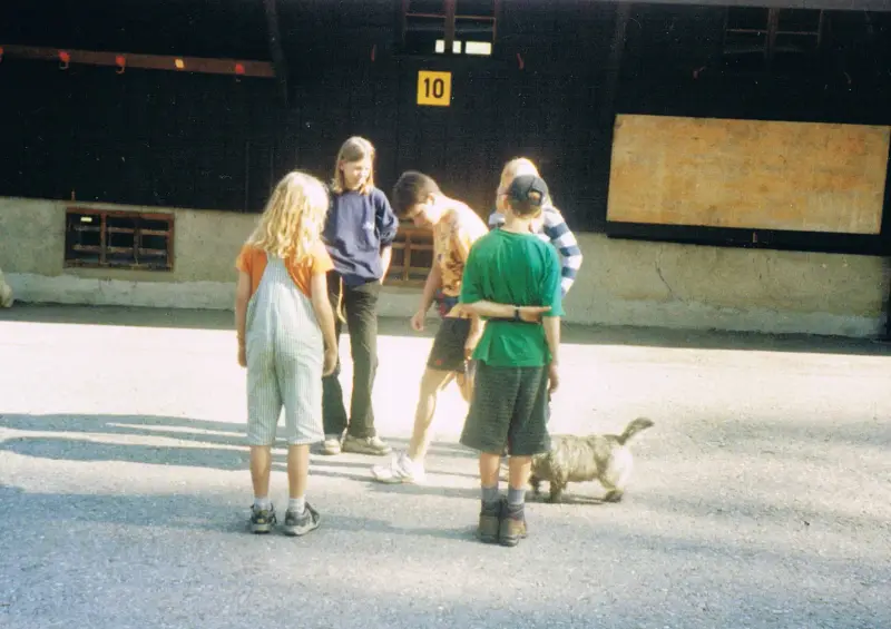 Eine Gruppe von Kindern, die vor einem Gebäude mit Fenstern und einem Brett mit Nummern stehen. Rasse: Dandie Dinmont Terrier. – Jugend und Hund Lager 2001 in Eigenthal