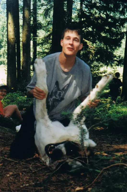 Das Bild zeigt einen Mann, der auf dem Boden im Wald sitzt und einen weissen und schwarzen Hund in den Händen hält. – Jugend und Hund Lager 2001 in Eigenthal