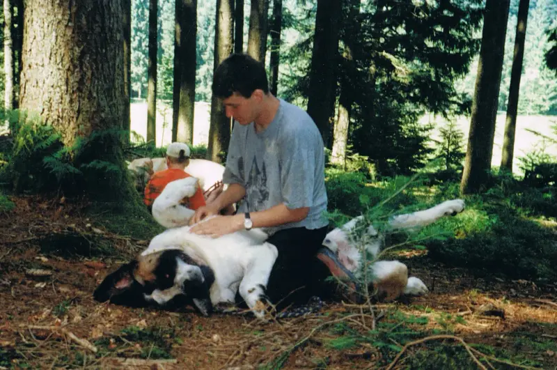 Das Bild zeigt einen Mann, der auf dem Boden neben einem grossen Hund sitzt, umgeben von Bäumen und Pflanzen. – Jugend und Hund Lager 2001 in Eigenthal
