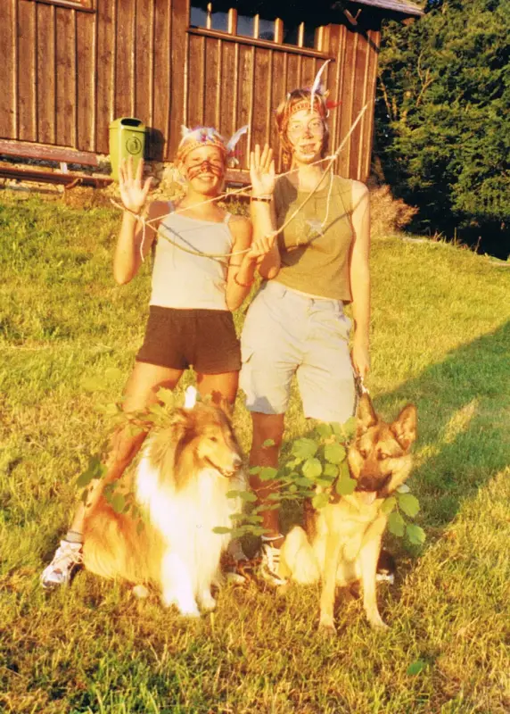 Zwei Menschen, die mit ihren Hunden im Gras stehen, umgeben von Bäumen und einem Holzhaus im Hintergrund. Rasse: Collie. – Jugend und Hund Lager 2001 in Eigenthal