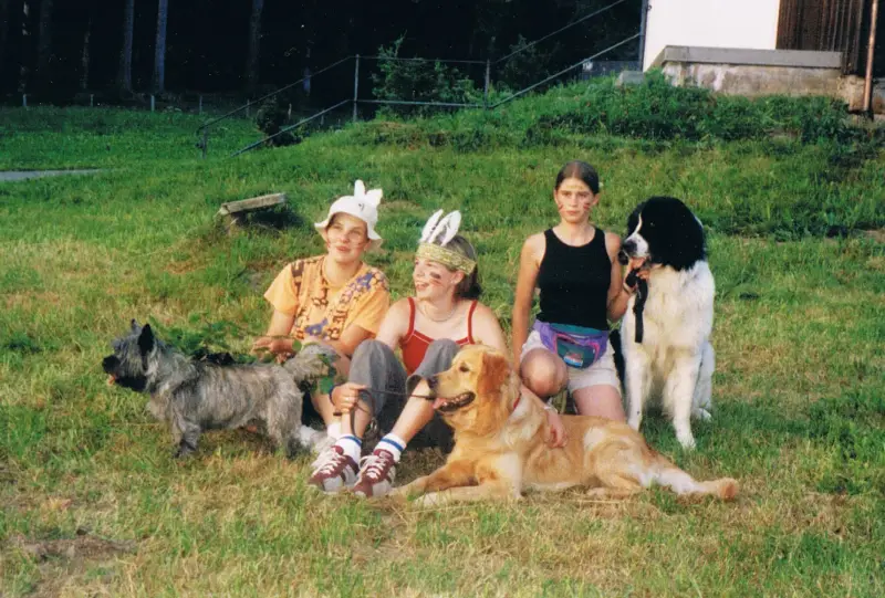 Eine Gruppe von vier Personen, die auf einem üppigen grünen Feld sitzen, wobei zwei von ihnen Mützen… Rassen: Chow-Chow, Cairn Terrier. – Jugend und Hund Lager 2001 in Eigenthal