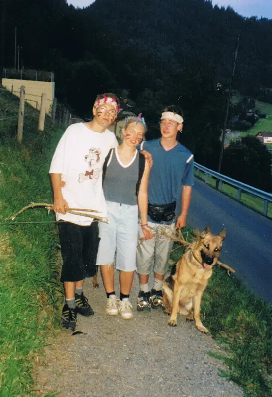 Drei Personen, die neben einem Hund auf einer Strasse stehen, die von Gras, einem Zaun, einer Mauer, einer… Rasse: Deutscher Schäferhund. – Jugend und Hund Lager 2001 in Eigenthal