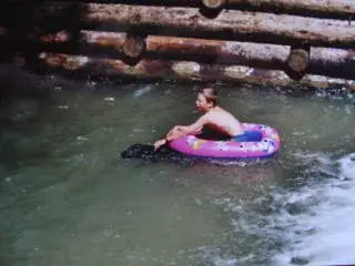 Das Bild zeigt einen Mann, der auf einem aufblasbaren Floss im Wasser schwebt, mit einer hölzernen Holzbalkenwand im Hintergrund. – Jugend und Hund Lager 2001 in Eigenthal