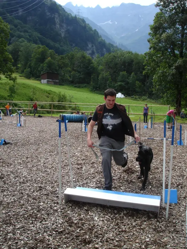 Das Bild zeigt einen Mann, der mit einem schwarzen Hund auf einer Leine in einem Feld läuft, umgeben von einer Gruppe von Menschen, einem… – Jugend und Hund Lager 2011 in Elm