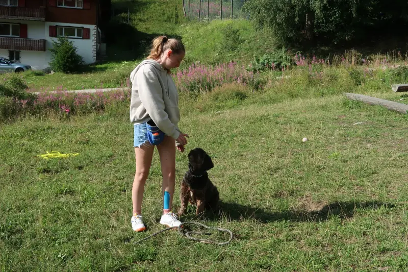 Eine Frau, die mit ihrem Hund im Gras steht, eine Jacke trägt und eine Leine hält. Rasse: Bouvier des Flandres. – Jugend und Hund Lager 2023 in Segnas