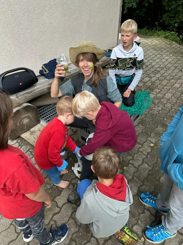 Eine Gruppe von Kindern, die um eine Frau sitzen und einen Hut tragen und eine Flasche in der Hand halten. – Jugend und Hund Lager 2025 in Segnas