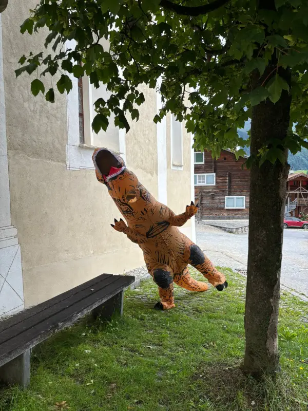 Eine Person in einem T-Rex Kostüm, die auf dem Gras vor einem Baum steht, mit einer Bank auf der linken Seite und einem Gebäude mit… – Jugend und Hund Lager 2025 in Segnas