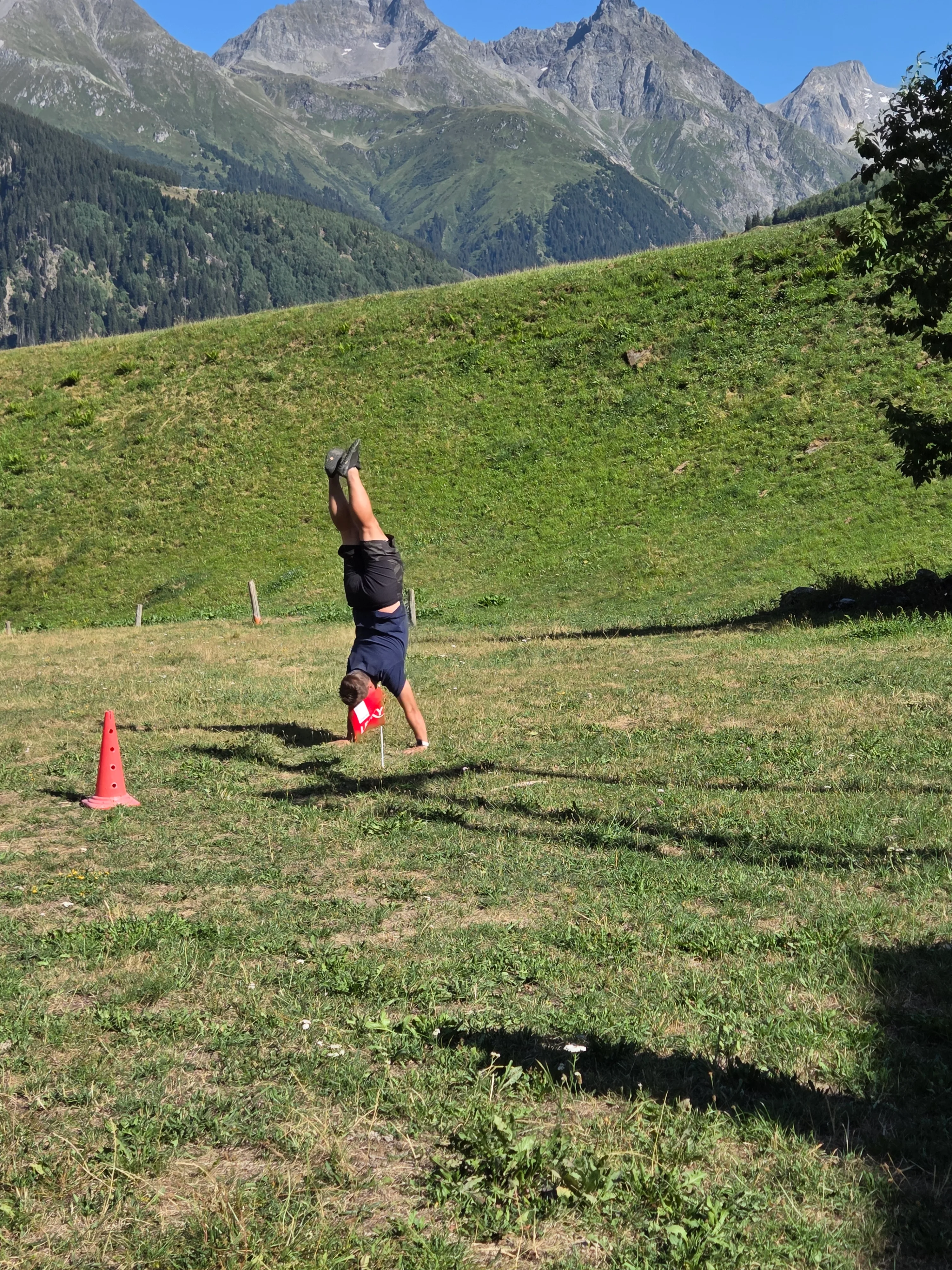 Das Bild zeigt einen Mann in einem schwarzen Hemd und schwarze Hosen, die auf einem üppigen grünen Hang stehen, mit einem roten Kegel vor ihm. Im Hintergrund gibt es Bäume, Berge und einen klaren blauen Himmel. Aktivität: Chillen (chill).