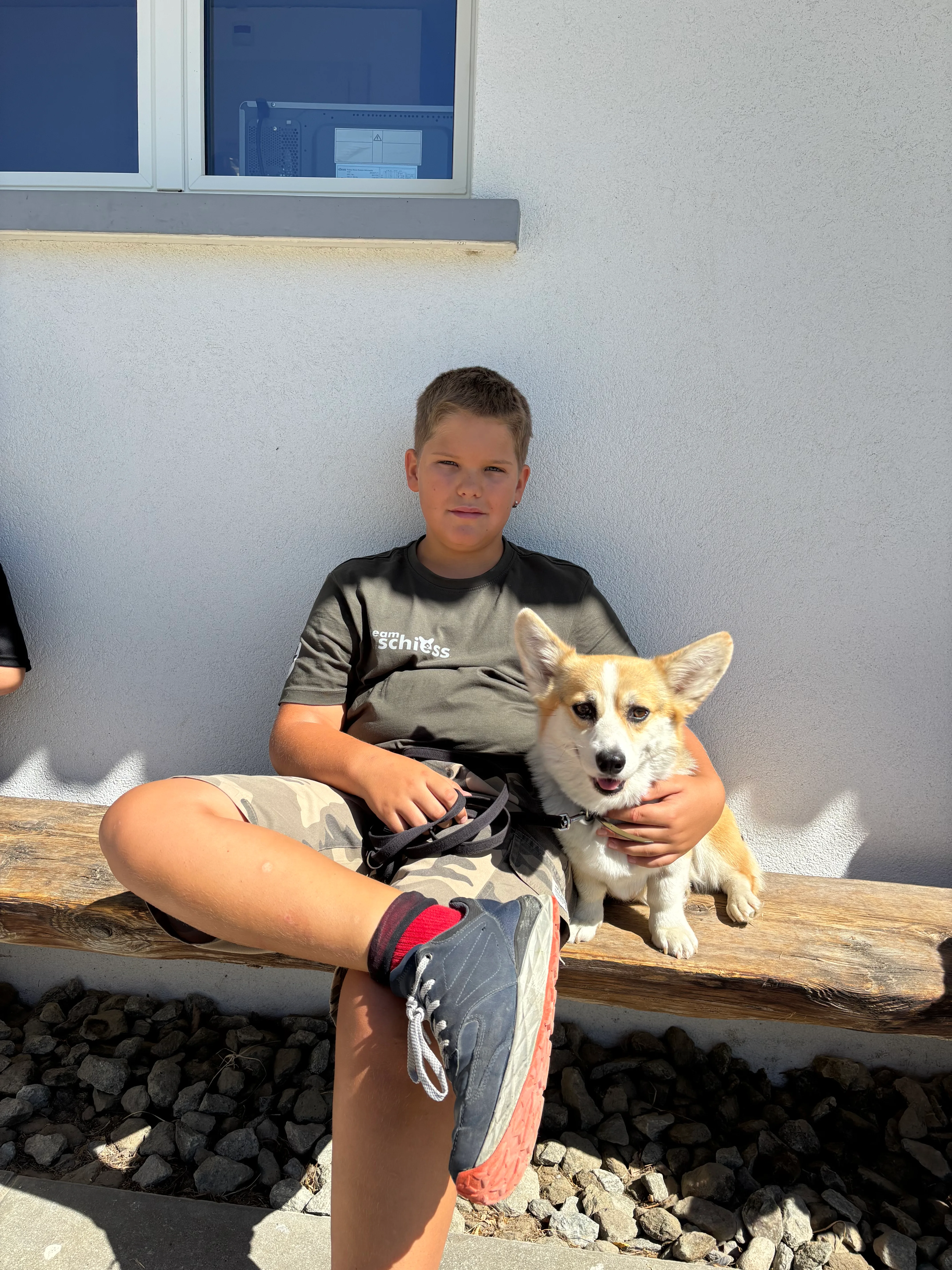 Das Bild zeigt einen jungen Jungen, der auf einer Holzbank sitzt, mit einem Hund in den Armen. Er trägt ein schwarzes T-Shirt und Shorts, und neben ihm befindet sich ein Paar Turnschuhe. Im Hintergrund sind Steine und eine Wand. Hunderassen: Hund. Aktivität: Wandern (hiking).