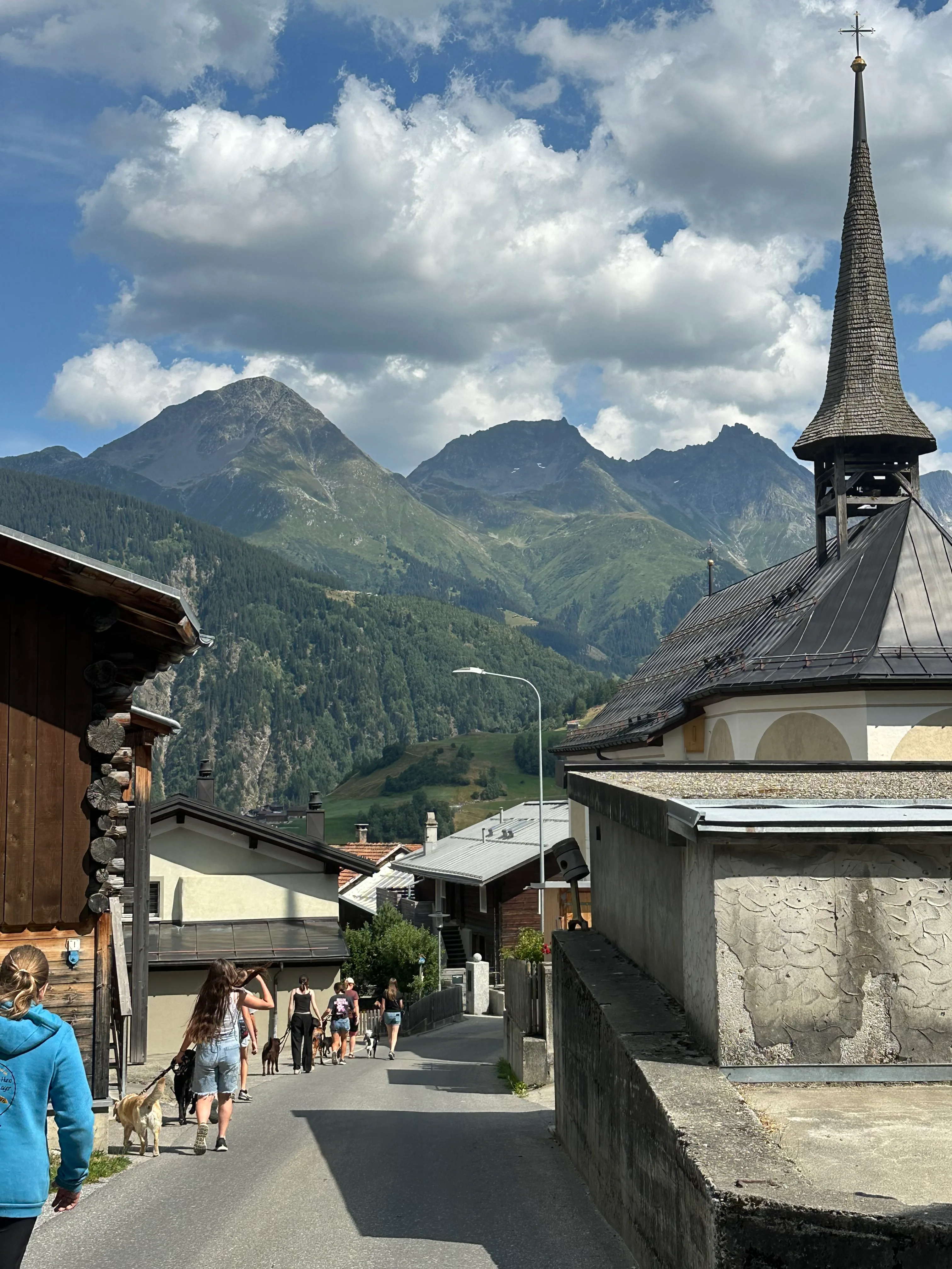 Das Bild zeigt eine Gruppe von Menschen, die auf der Seite einer Straße neben einer Kirche stehen, mit einer Person, die eine blaue Jacke trägt und eine Handtasche trägt. Im Hintergrund befinden sich Gebäude, Bäume, Berge, Wolken und ein klarer blauer Himmel. Hunderassen: Hund. Aktivität: Chillen (chill).