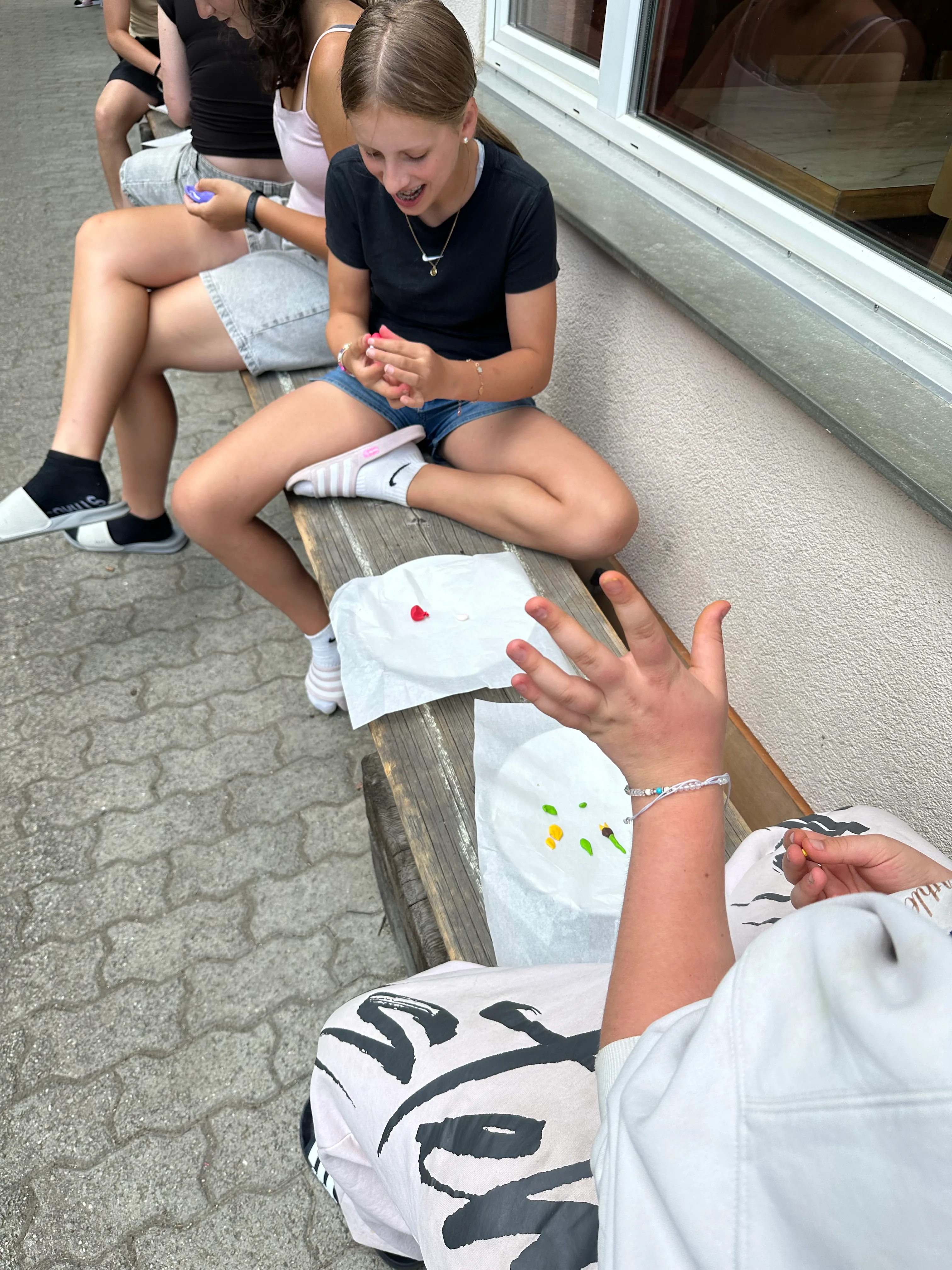 Das Bild zeigt eine Gruppe von Menschen, die auf einer Holzbank sitzen. Auf der rechten Seite der Bank steht eine Person, die ein Papier in der Hand hält. Im Hintergrund befindet sich eine Wand und ein Glasfenster. Aktivität: Wandern (hiking).