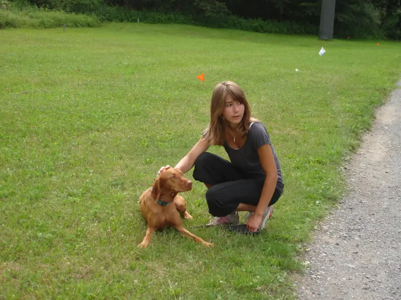Eine Frau, die neben einem braunen Vizsla-Hund auf einem Grasfeld kniet. Rasse: Vizsla. – Jugend und Hund Lager 2009 in Ruen