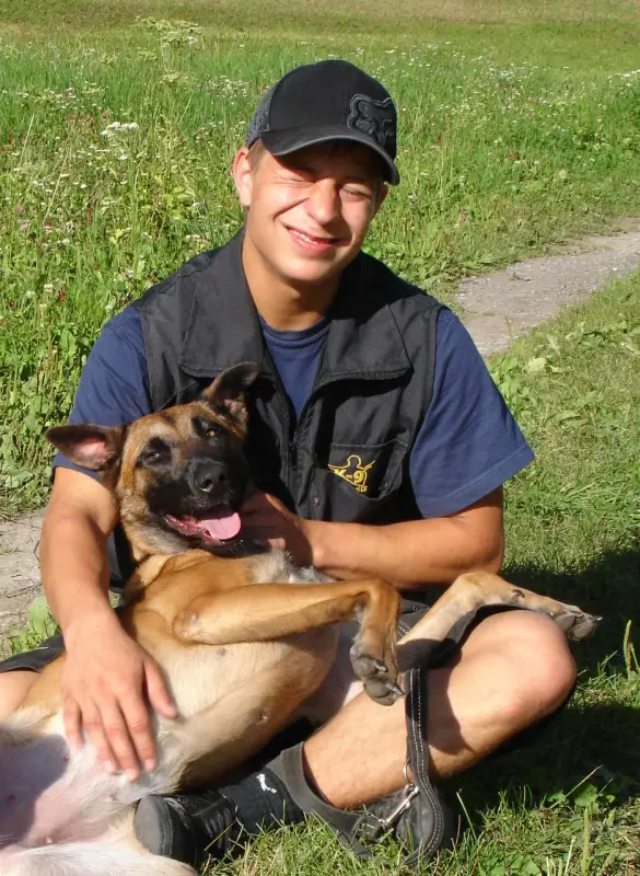 Das Bild zeigt einen Mann mit einer Mütze und einem blauen T-Shirt, der mit einem Hund in den Armen auf dem Gras sitzt. Rasse: Malinois. – Jugend und Hund Lager 2009 in Ruen