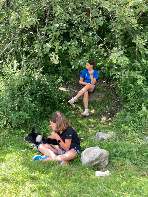 Zwei Frauen, die mit ihrem Hund auf dem Gras sitzen, umgeben von Steinen und Bäumen im Hintergrund. – Jugend und Hund Lager 2023 in Segnas