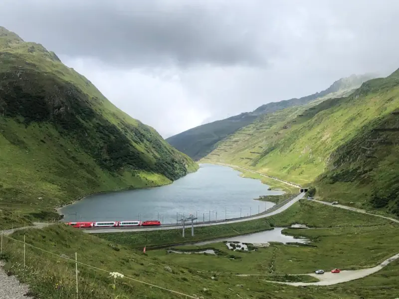 Das Bild zeigt einen Zug, der eine kurvenreiche Strasse neben einem Gewässer hinunterfährt, umgeben von üppig grünen Hügeln und einem… – Jugend und Hund Lager 2023 in Segnas
