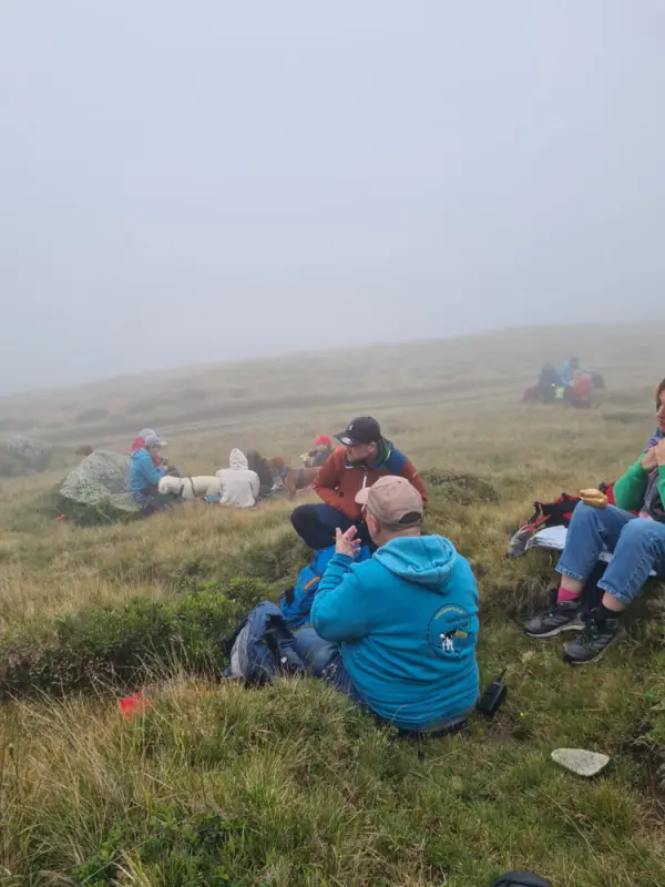 Eine Gruppe von Menschen, die auf einem grasbedeckten Hang sitzen, mit Taschen und anderen Gegenständen, die um sie herum verstreut sind. – Jugend und Hund Lager 2023 in Segnas