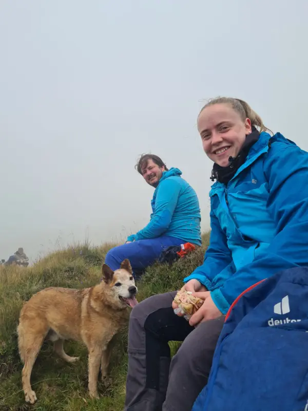 Das Bild zeigt einen Mann und eine Frau, die auf einem grasbedeckten Hang sitzen und sowohl blaue Jacken… Rasse: Australian Catlle Dog. – Jugend und Hund Lager 2023 in Segnas