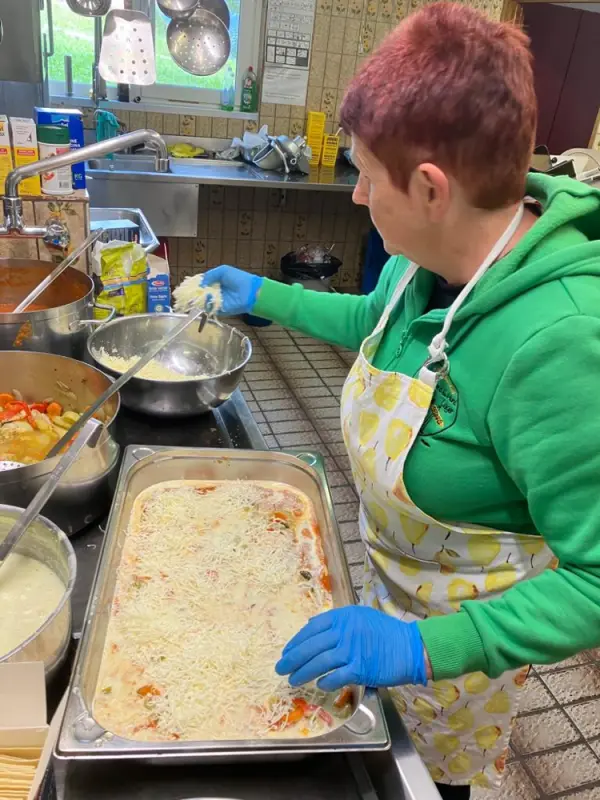 Eine Frau in einer Schürze und blaue Handschuhe, die Essen in einer Küche zubereiten. – Jugend und Hund Lager 2023 in Segnas