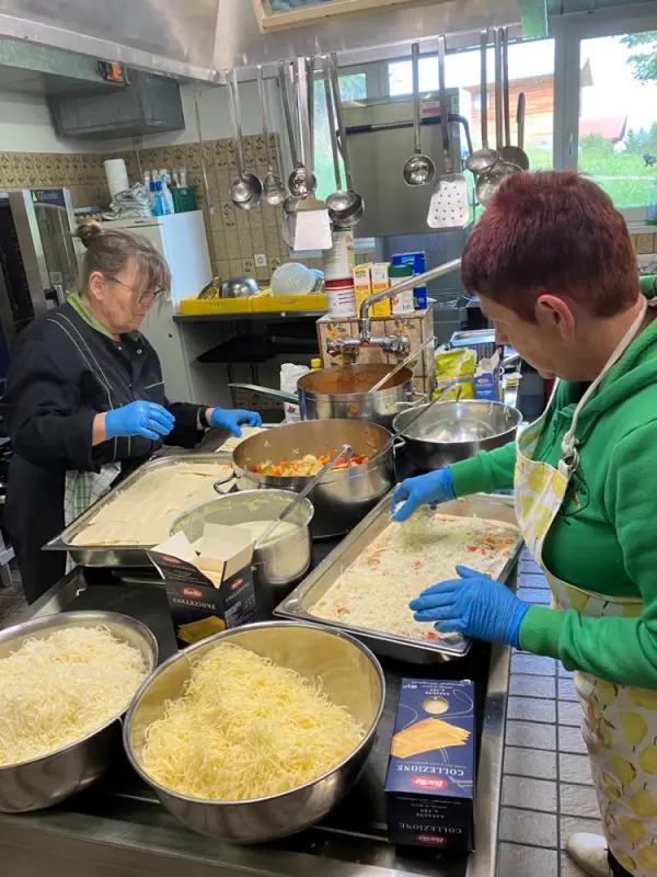 Zwei Frauen in Schürzen und blaue Handschuhe, die Essen in einer Küche zubereiten. – Jugend und Hund Lager 2023 in Segnas