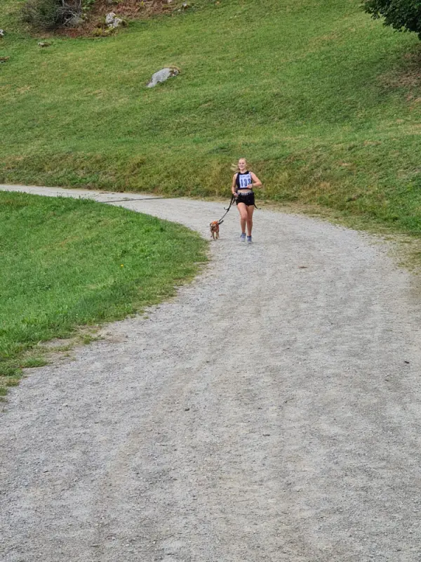 Eine Frau, die mit ihrem Hund eine unbefestigte Strasse hinunterläuft, umgeben von üppig grünem Gras und Bäumen auf beiden Seiten. – Jugend und Hund Lager 2023 in Segnas