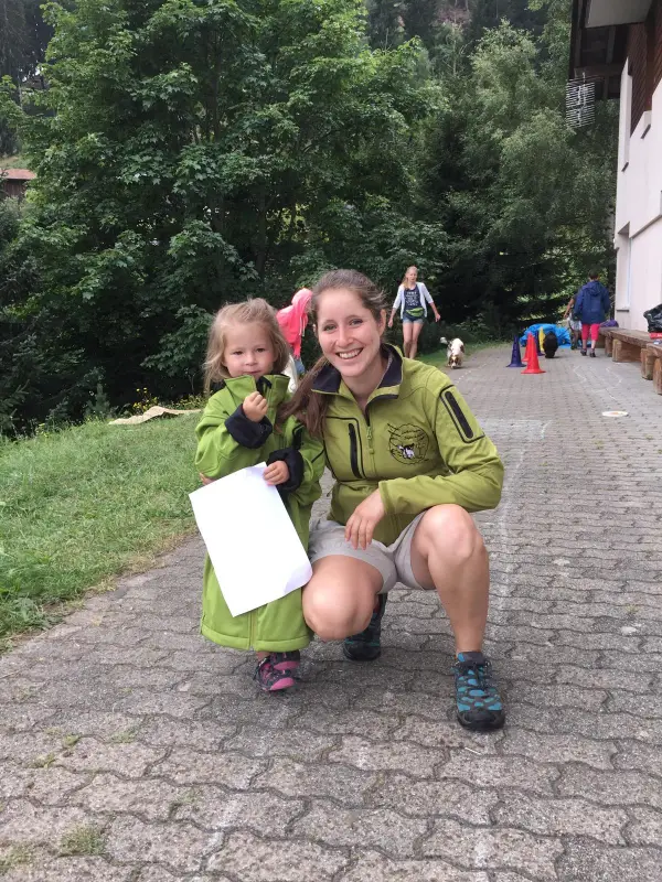 Ein junges Mädchen in einer grünen Jacke und blaue Schuhe hocken auf einem kleinen Kind in einer grünen Jacke. – Jugend und Hund Lager 2024 in Segnas
