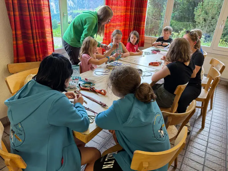 Eine Gruppe von Kindern, die um einen Tisch sitzen, in einem Raum mit einer mit Vorhängen und Glasfenstern geschmückten Wand, durch die… – Jugend und Hund Lager 2025 in Segnas