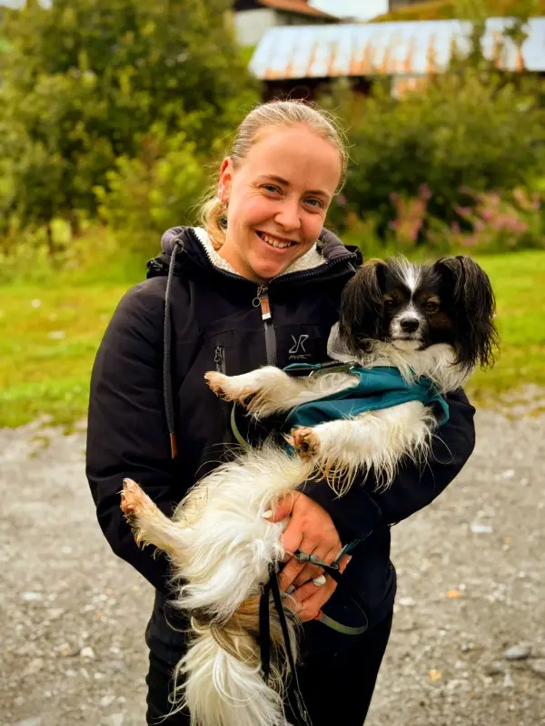 Eine Frau, die auf einer Strasse steht, lächelnd und einen kleinen Hund in den Armen hält. – Jugend und Hund Lager 2025 in Segnas
