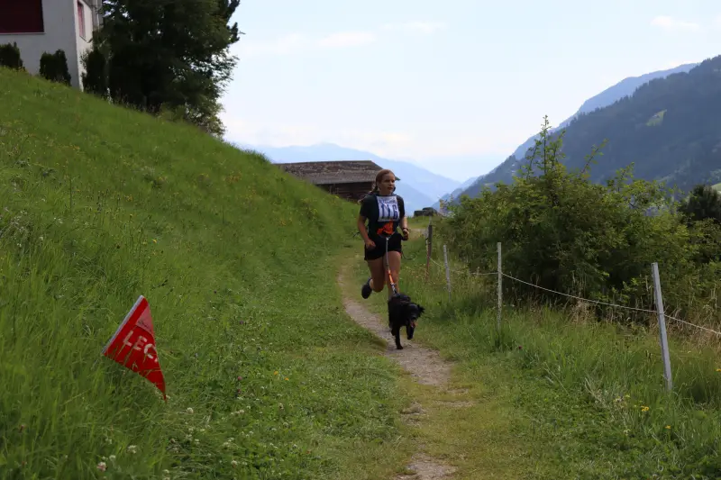 Eine Frau, die eine unbefestigte Strasse mit einem Hund hinunterläuft, umgeben von Gras, Pflanzen, Blumen, einem Zaun, einem Haus mit einem… – Jugend und Hund Lager 2025 in Segnas