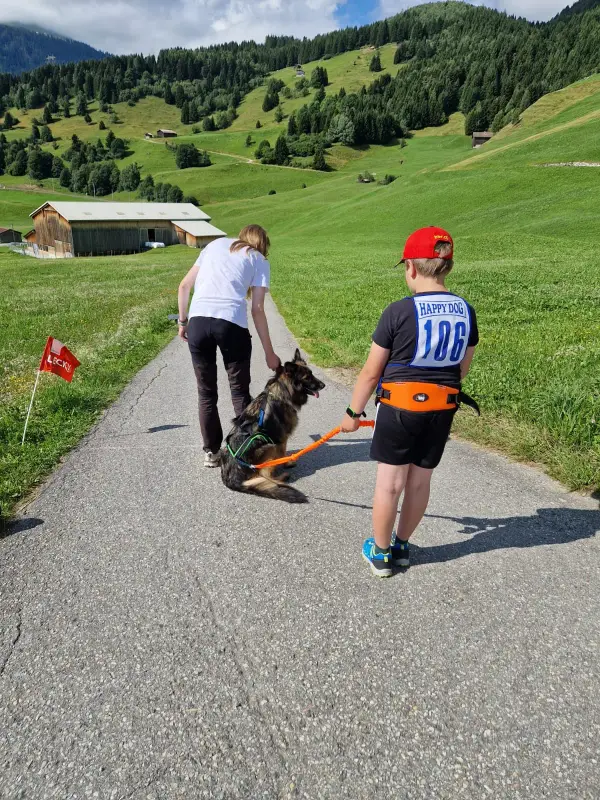 Eine Frau und einen Jungen, die ihren Hund eine Strasse entlang laufen, umgeben von üppigem grünem Gras, Pflanzen und Bäumen. – Jugend und Hund Lager 2025 in Segnas
