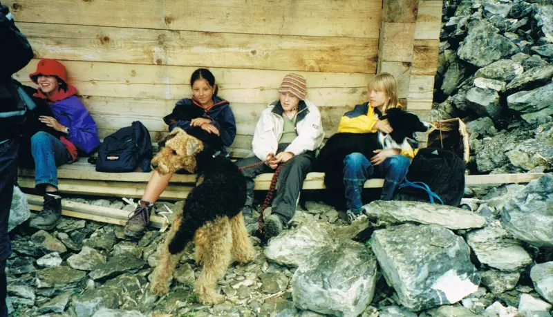 Eine Gruppe von Menschen, die auf einer Holzbank sitzen, mit einem schwarzen und braunen Airedale Terrier im Vordergrund. – Jugend und Hund Lager 2000 in Sörenberg