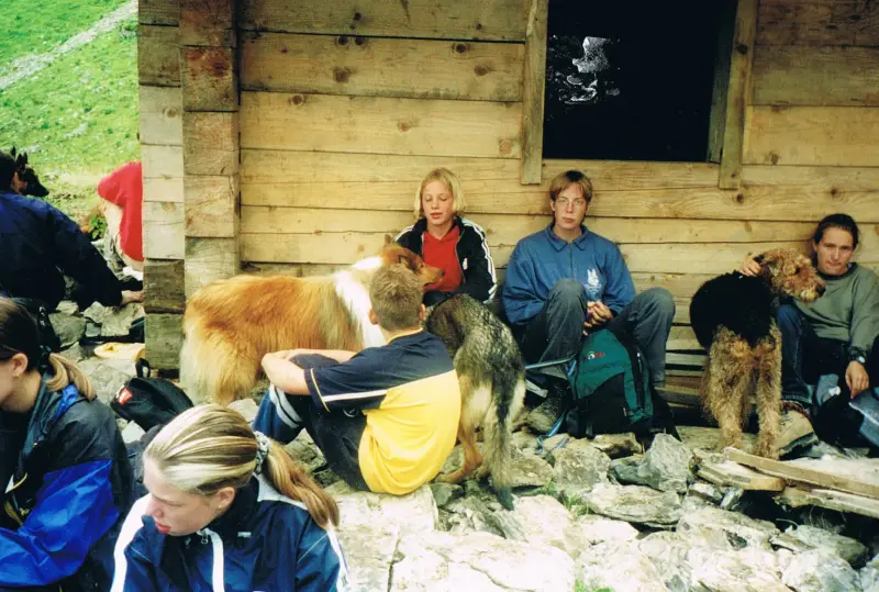 Eine Gruppe von Menschen, die auf dem Boden vor einem Holzhaus sitzen, mit ein paar Hunden im Vordergrund. – Jugend und Hund Lager 2000 in Sörenberg