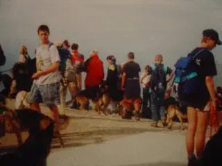 Eine Gruppe von Menschen, die mit ihren Hunden an einem Strand spazieren, mit dem Himmel im Hintergrund. – Jugend und Hund Lager 2000 in Sörenberg