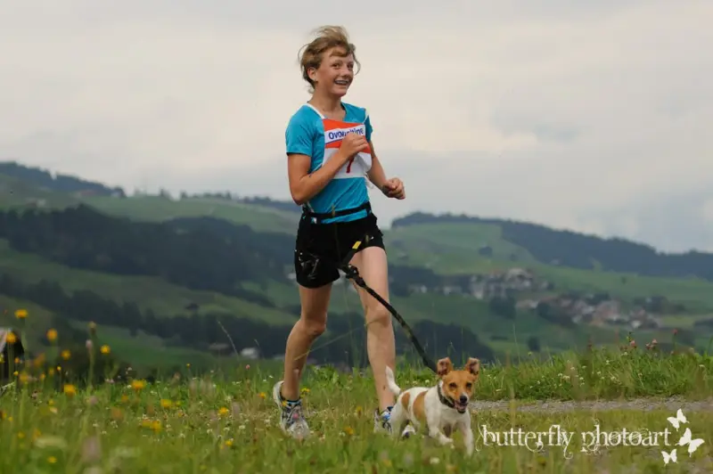 Eine Frau, die mit ihrem Hund an einer Leine in einem Feld läuft, umgeben von Pflanzen mit Blumen, Bäumen, Hügeln und… Rasse: Toy Terrier. – Jugend und Hund Lager 2012 in Vignogn