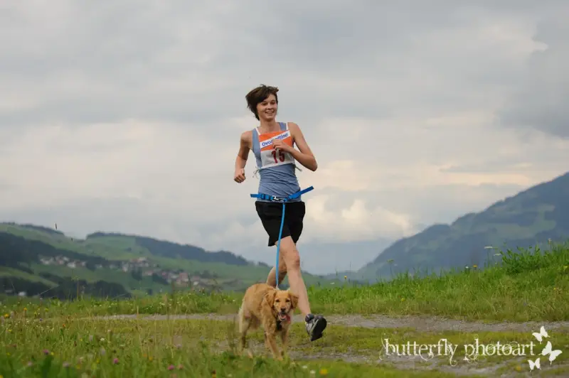 Eine Frau, die mit ihrem Hund auf einem Feld läuft, umgeben von üppig grünem Gras, lebendigen Blumen, hohen Bäumen, hügeligen Hügeln und… – Jugend und Hund Lager 2012 in Vignogn